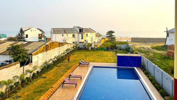 A swimming pool with lounge chairs, a grassy area, and houses in the background. - Tiger Inn Beach Resort (Mandarmani)