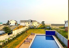 A swimming pool with lounge chairs, a grassy area, and houses in the background. - Tiger Inn Beach Resort (Mandarmani)