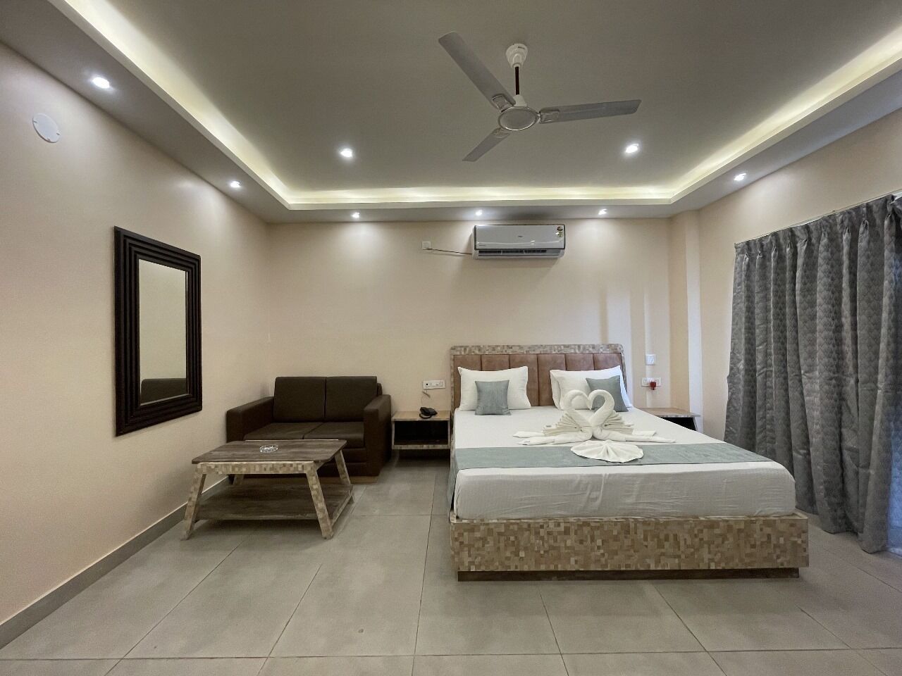 A modern hotel room with a large bed, a ceiling fan, recessed lighting, a sofa, a small table, and a wall-mounted air conditioner.
