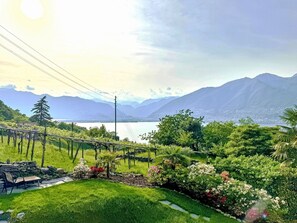 A garden with a bench, a stone wall, and a view of mountains and a lake. - Casa Sole by Interhome (Magadino)
