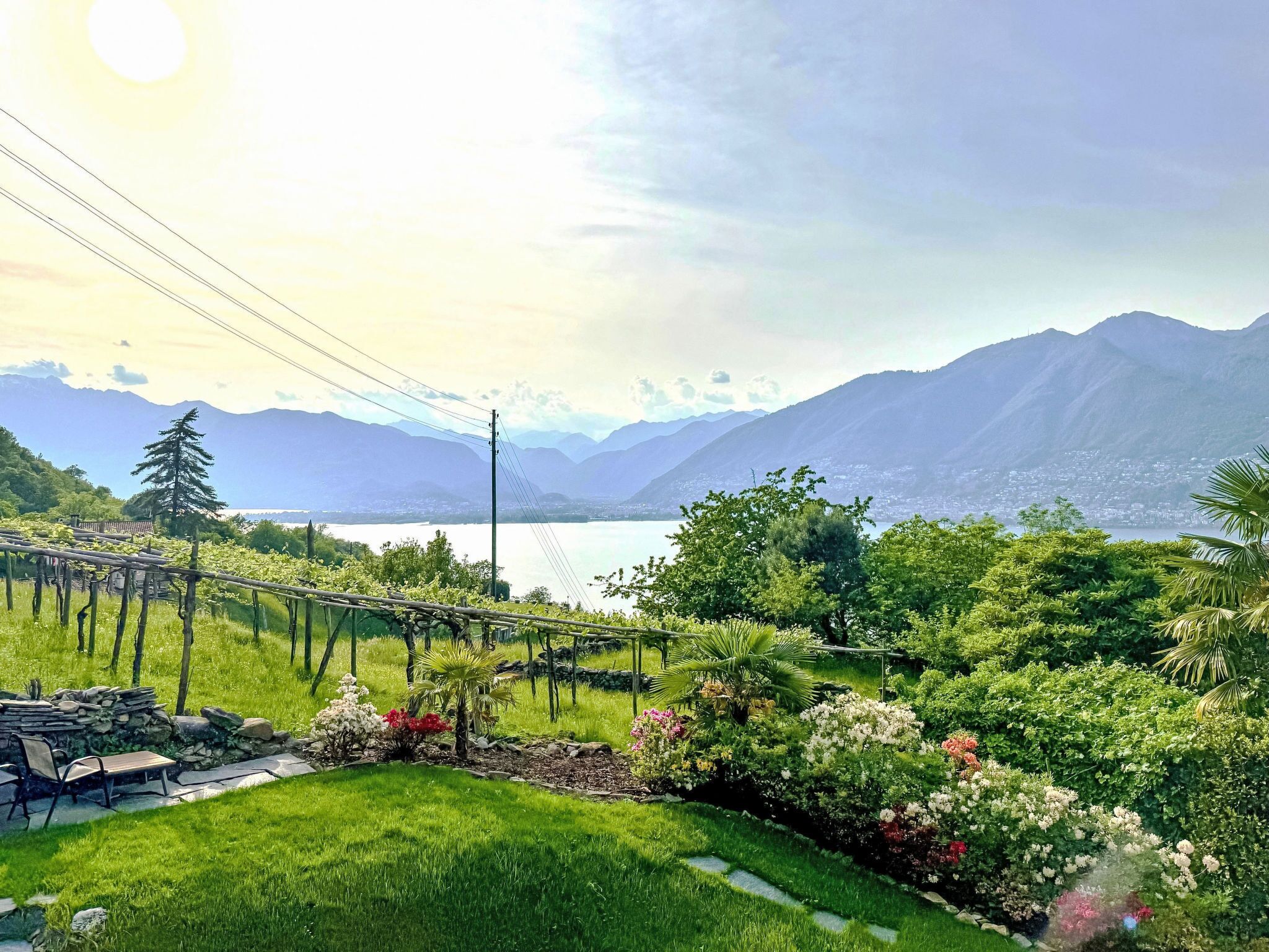 A lush garden with a bench, a stone wall, and a view of mountains and a lake.