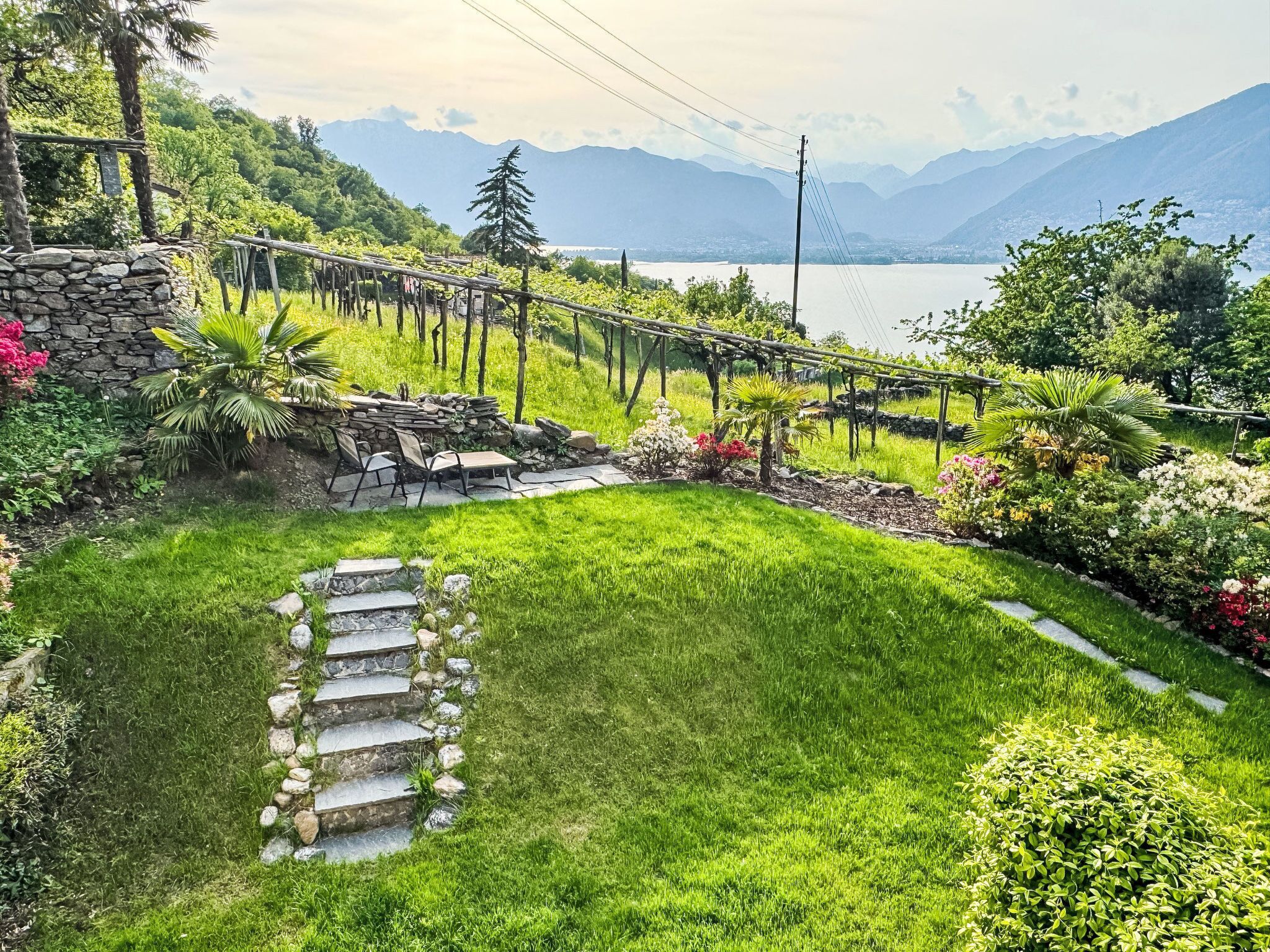 A garden with a stone staircase leading to a seating area, surrounded by lush greenery and a view of mountains and a lake.