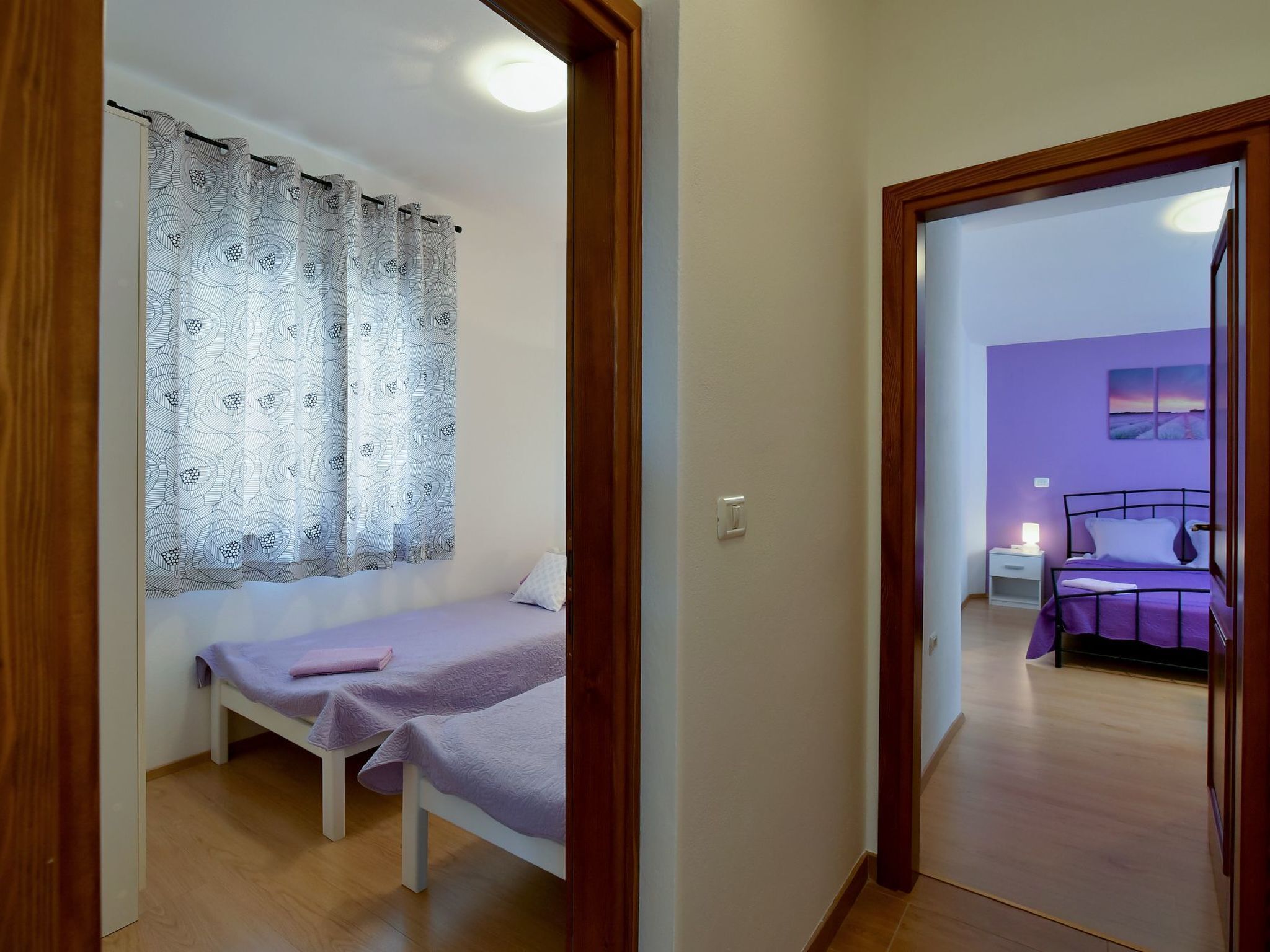 Une chambre avec deux lits simples, un rideau à motifs et une vue sur une autre pièce dont le mur est peint en violet et où se trouve un lit.