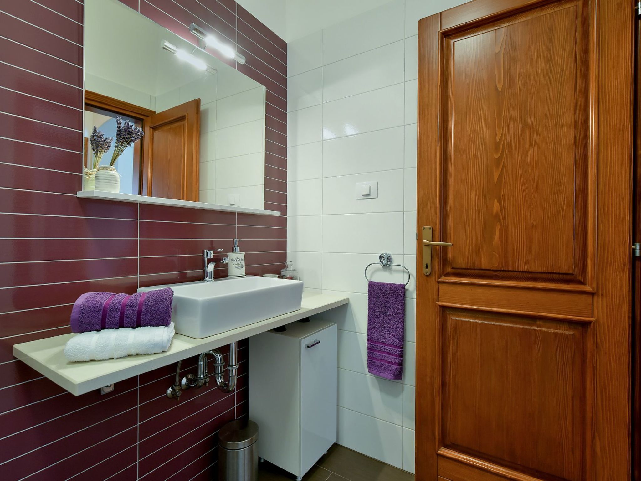Une salle de bain avec une porte en bois, un lavabo blanc avec un miroir au-dessus, et une étagère avec des serviettes.
