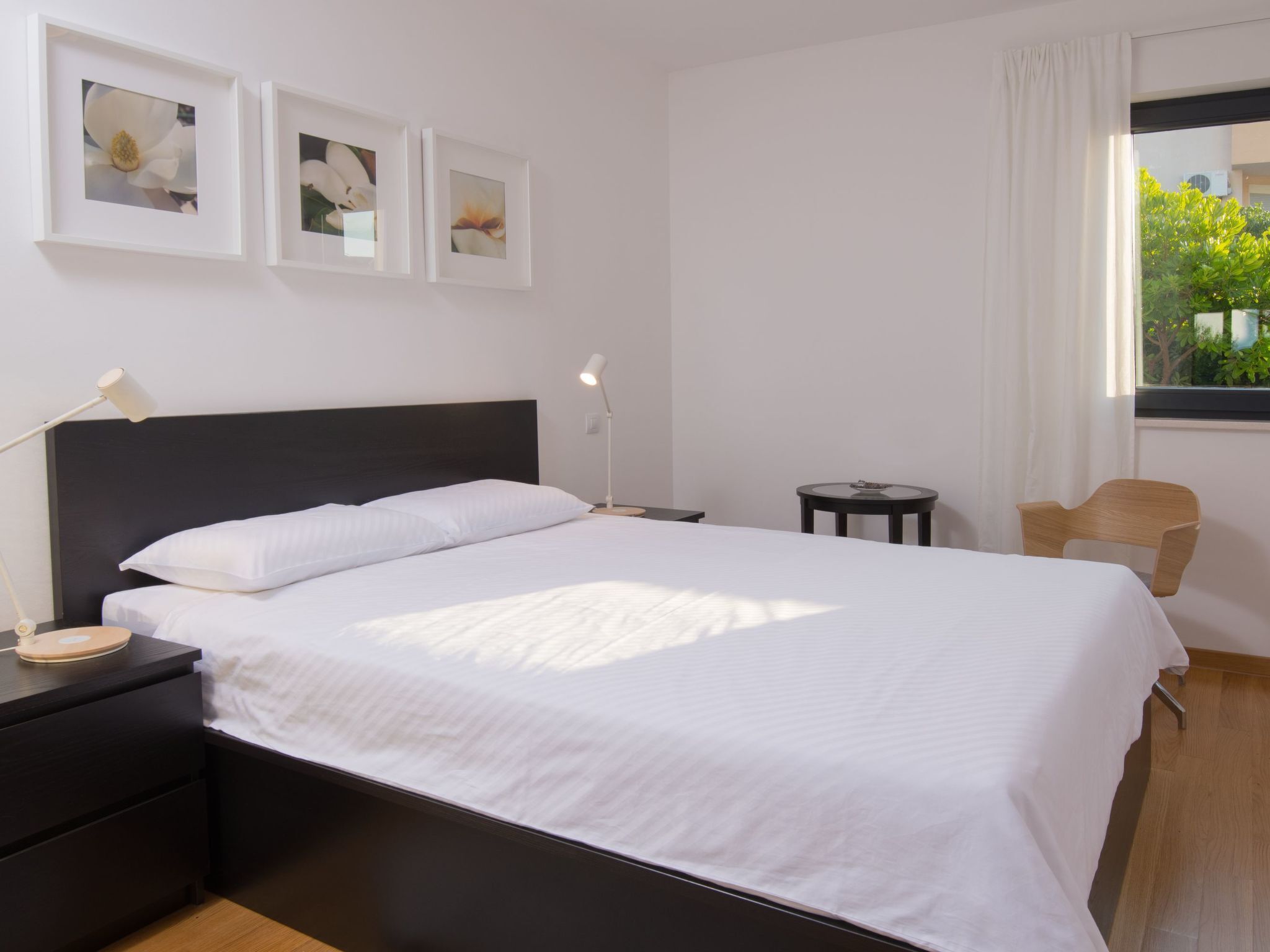 A bedroom with a black headboard, white bedding, a bedside table with a lamp, a small round table, a chair, and three framed pictures on the wall.