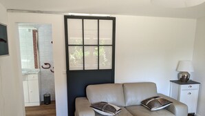 Een woonkamer met een beige bank, een klein rond tafeltje, een lamp en uitzicht op een tuin door een raam.