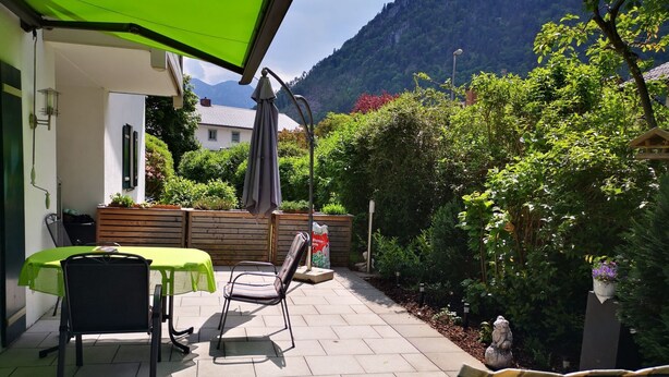 En terrasse med et bord og stole, en grøn parasol og udsigt til et bjerg.