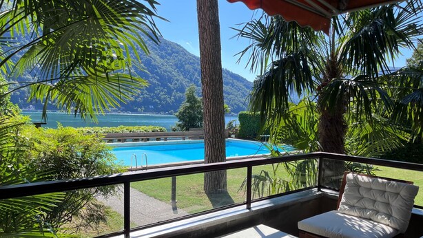 A balcony with a view of a swimming pool, surrounded by lush greenery and a mountain backdrop. - "Lake Escape", mit Pool by Interhome (MAROGGIA)