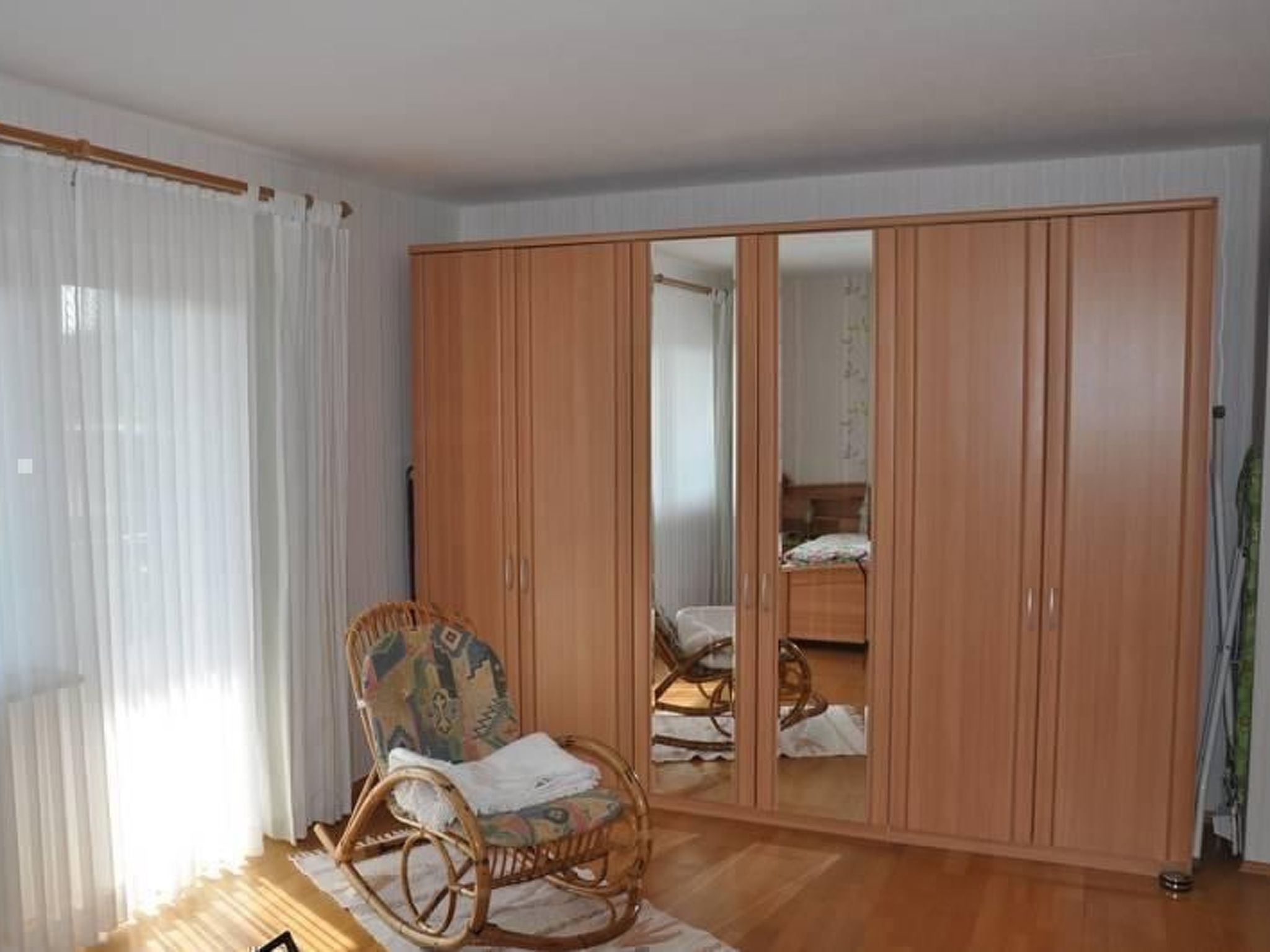 Une pièce comprenant une armoire en bois, un fauteuil à bascule en osier et un lit recouvert d’une courtepointe blanche.