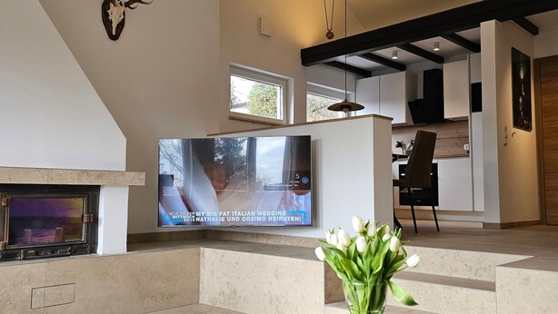 Una sala de estar moderna con chimenea, un televisor de pantalla plana montado en la pared y un florero con flores blancas sobre una mesa redonda.