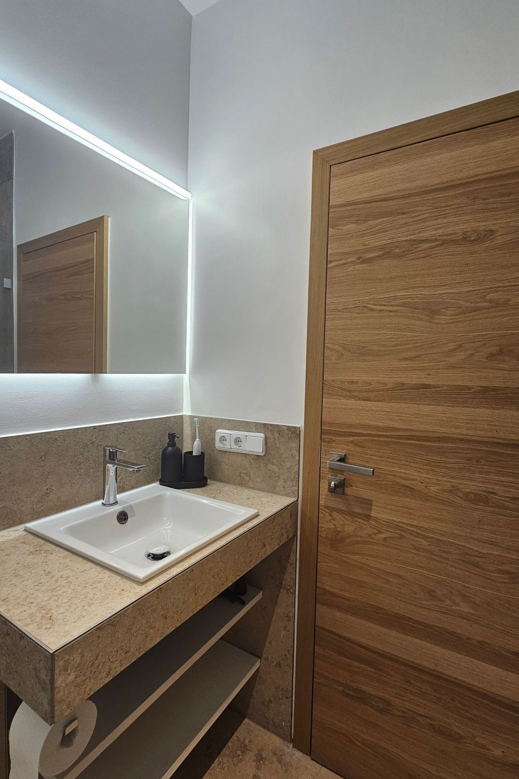 Ein Badezimmer mit Holztür, weißem Waschbecken, Spiegel mit Unterschrankbeleuchtung und Handtuchhalter.