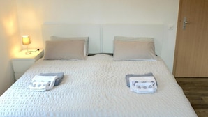 Uma cama de casal com um edredom branco texturizado, duas almofadas e toalhas dobradas sobre a cama.