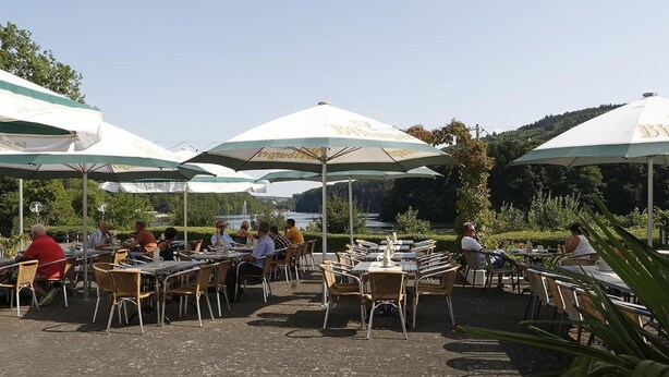 Un espace de restauration en plein air, avec des tables et des chaises placées sous des parasols, offrant une vue sur une étendue d’eau.