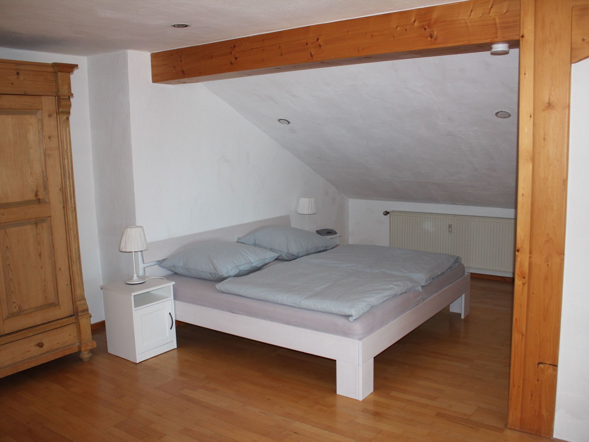 A bedroom with a sloped ceiling, a wooden wardrobe, a bedside table with a lamp, and a bed with pillows.