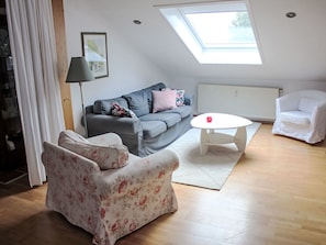 Een moderne woonkamer met een grijze bank, een fauteuil met bloemenprint, een witte salontafel en een witte fauteuil. Een dakraam zorgt voor natuurlijk licht.