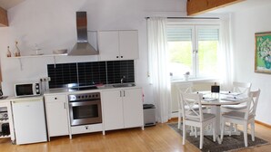 Une cuisine compacte avec des meubles blancs, un four à micro-ondes, une cuisinière et un évier. On y trouve également un coin repas avec une table et des chaises, ainsi qu’une fenêtre avec des rideaux.