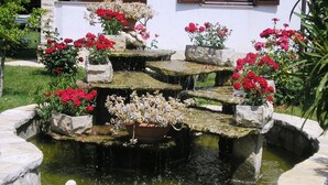 Una fuente de piedra con agua cayendo en cascada sobre los escalones, rodeada de macetas con flores rojas, frente a un edificio blanco con ventanas.
