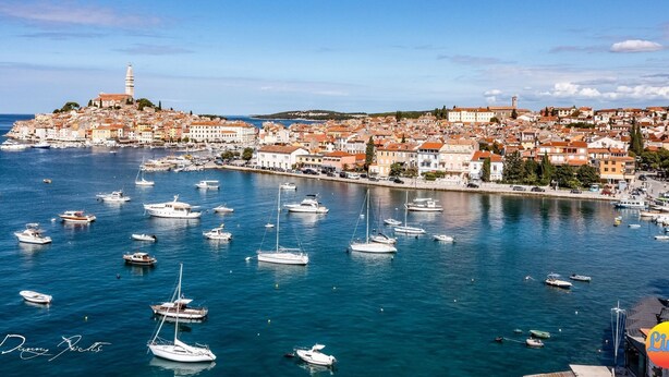 Un pueblo costero con una torre destacada, numerosos barcos en el puerto y una mezcla de edificios históricos y modernos.