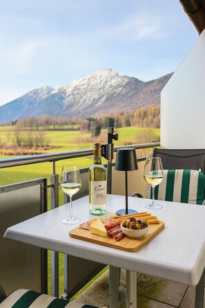 A balcony with a table set for a meal, featuring a bottle of Cades wine, glasses of white wine, a wooden board with cheese and cured meats, and a view of mountains. - Vacation apartment Anton in the vacation home Bayerisch Gmain, 68sqm, for 1 - 4 persons with 2 bedrooms and balcony (Bayerisch Gmain)