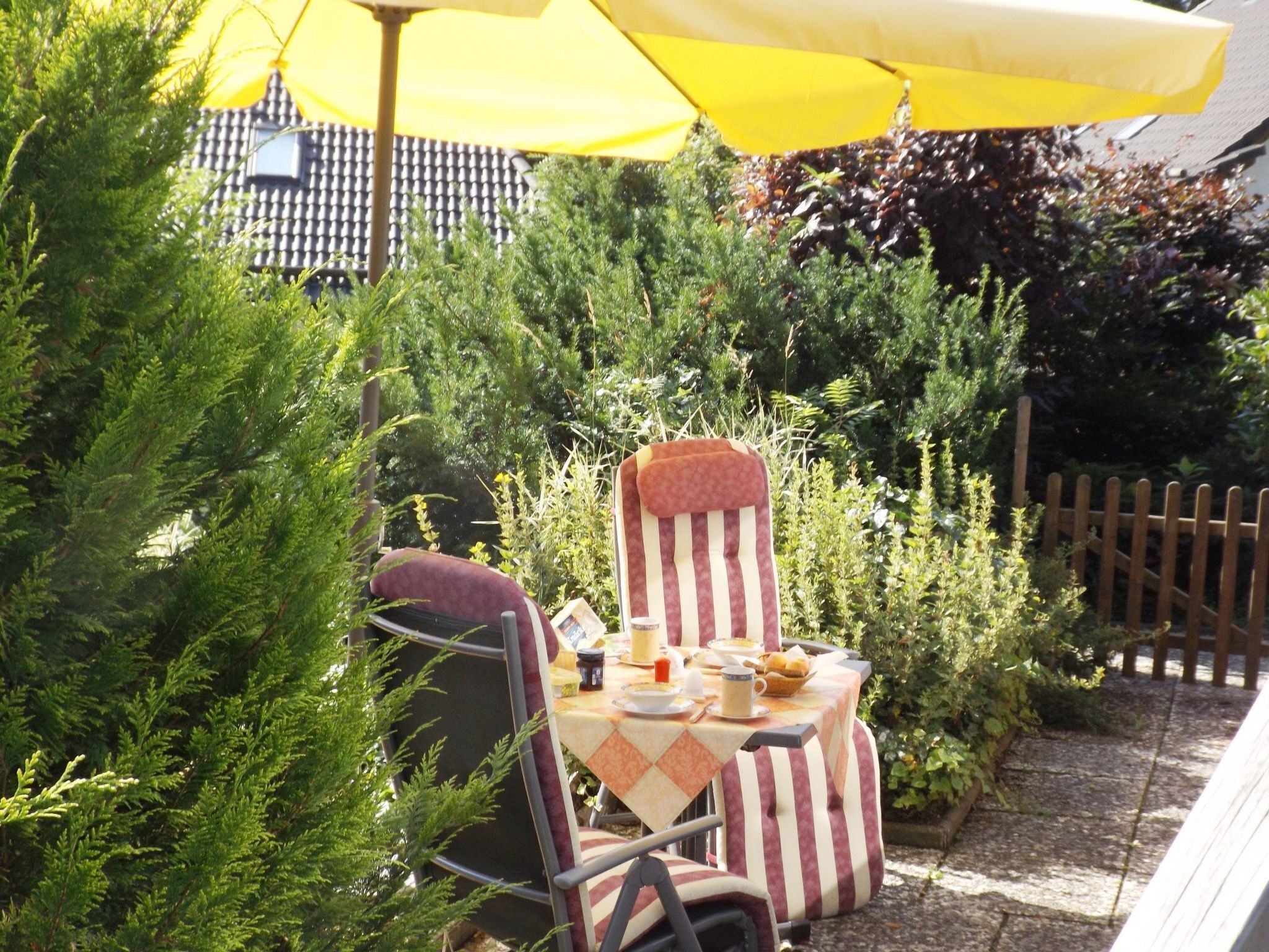 Eine Terrasse mit Tisch für zwei Personen, umgeben von Grünpflanzen und einem Holzzaun.