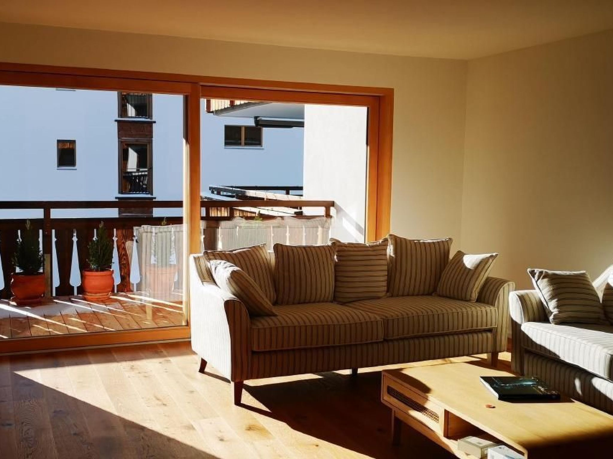 Una sala de estar moderna con un sofá y sillones, una mesa de centro de madera y un ventanal corredero que da a un balcón con plantas en macetas.