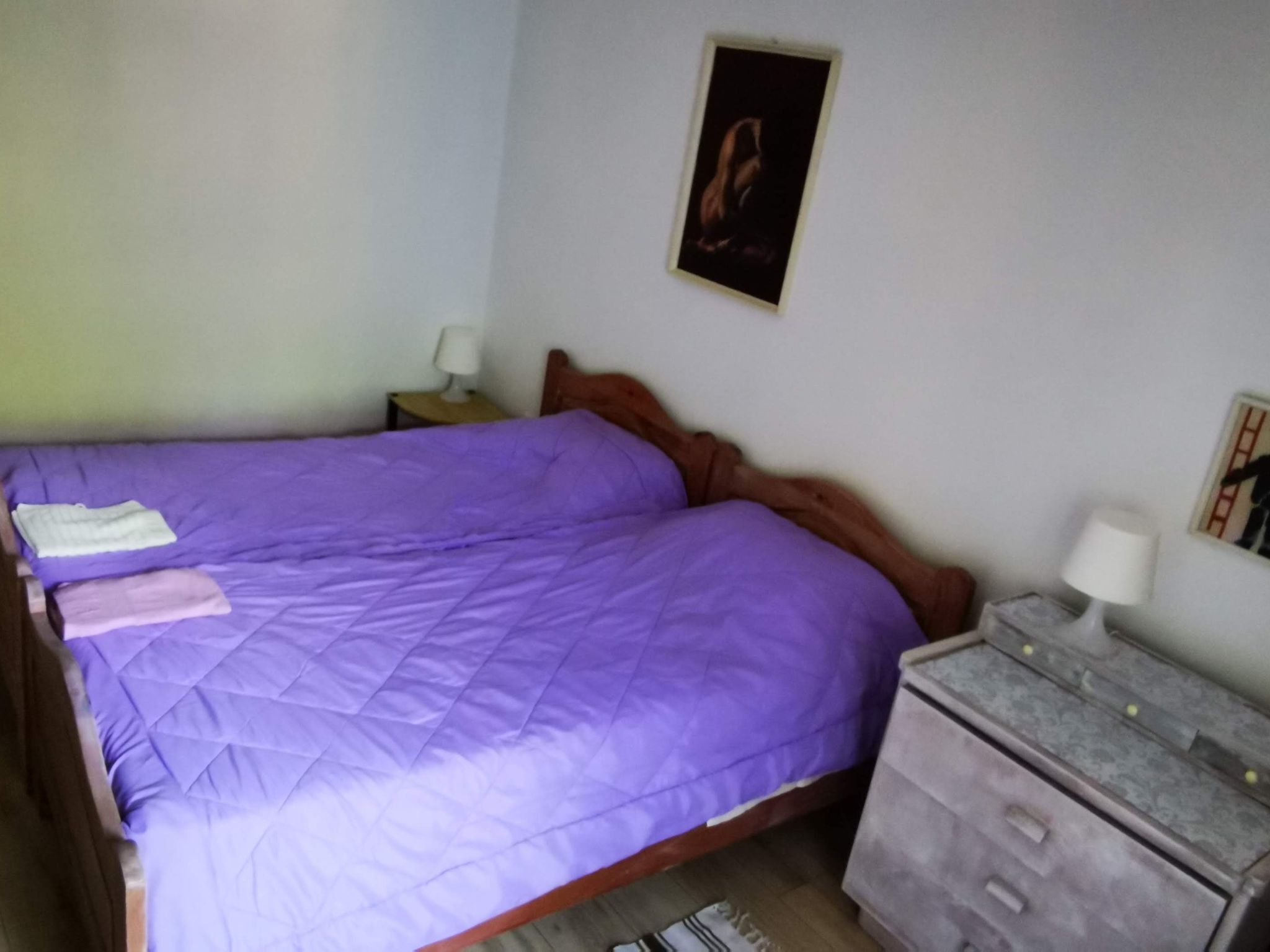 A bedroom with a purple bedspread, a wooden headboard, two bedside tables with lamps, and a framed picture on the wall.