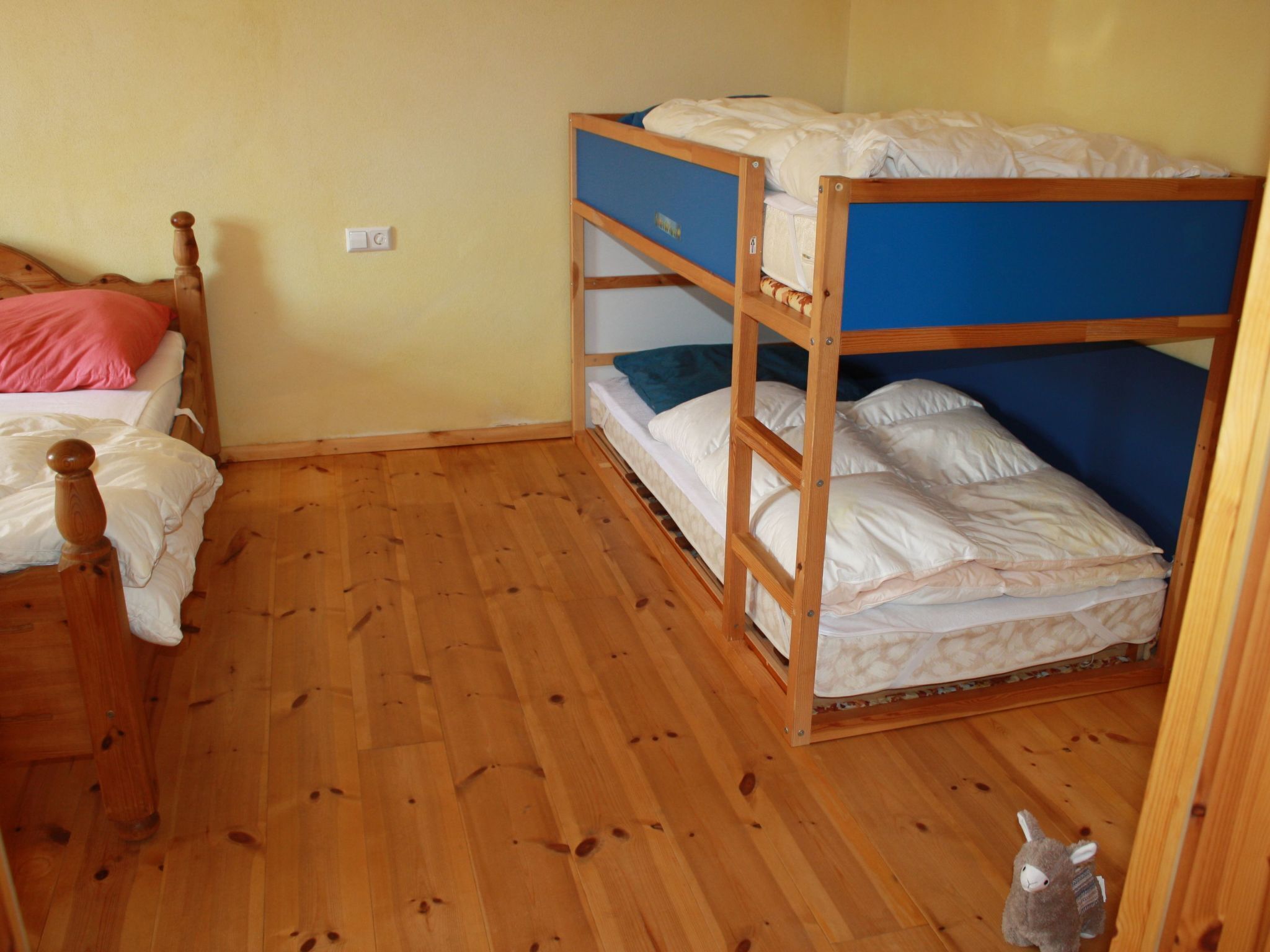 Une pièce avec un plancher en bois, un lit superposé avec une literie bleue et blanche, un lit simple avec une literie rose, et un petit animal en peluche sur le sol.