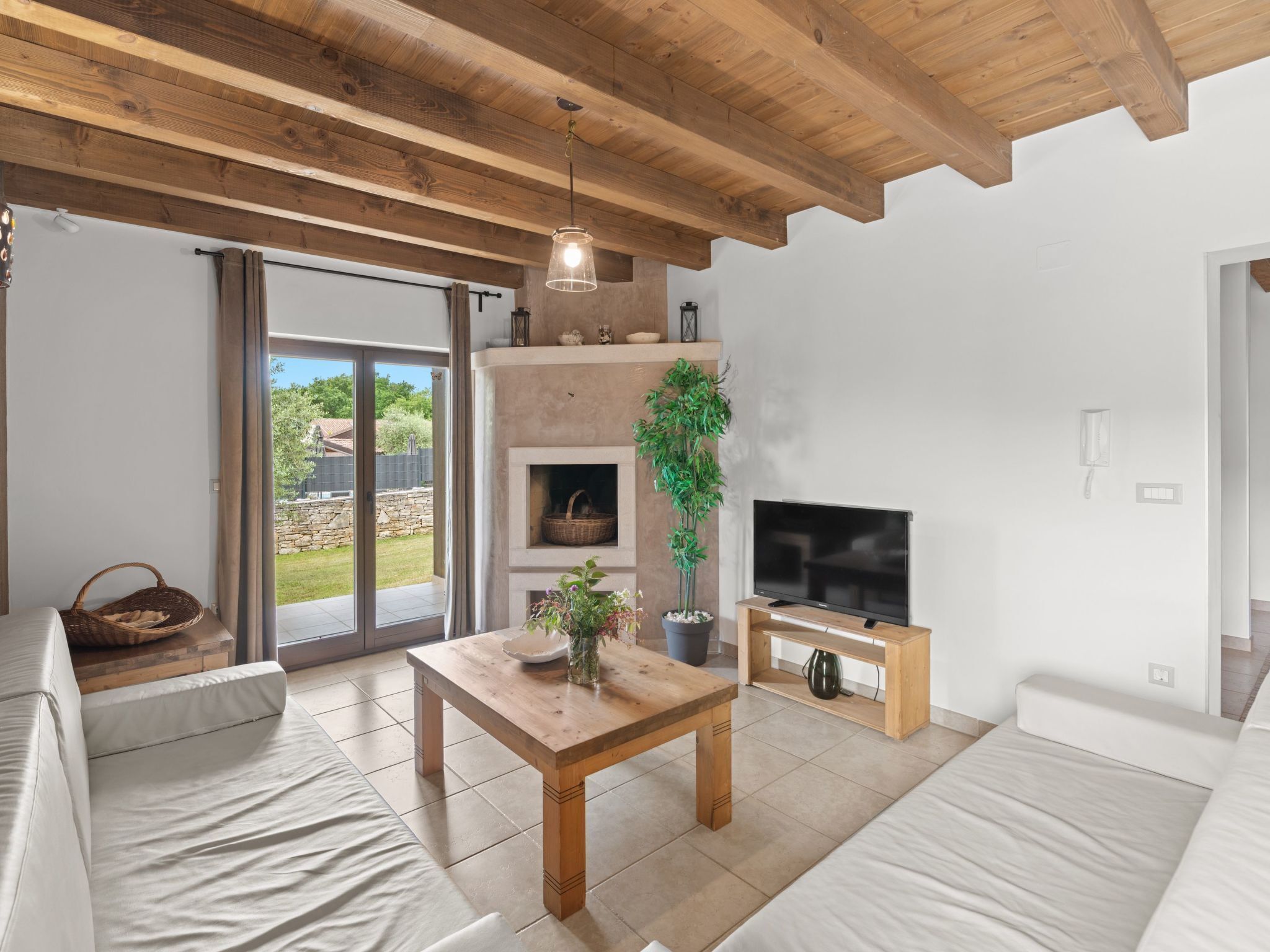A modern living room with a wooden ceiling, a fireplace, a television, and a large window overlooking a garden.