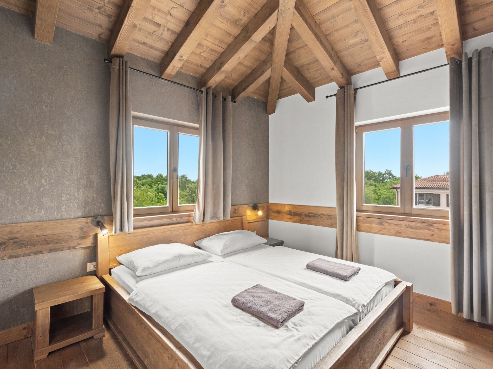 A bedroom with a wooden ceiling, two windows, a bed with white linens, and a nightstand.