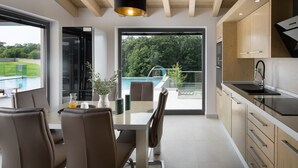 Una cuina moderna amb bigues de fusta, una zona de menjador amb taula de vidre, i vista a una piscina i vegetació exterior.
