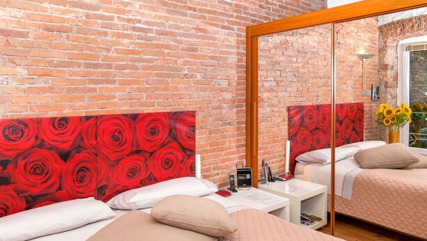Une chambre d’hôtel avec un mur en briques, une grande fresque représentant une rose rouge, un lit avec une literie beige, une table de chevet avec une lampe et une fenêtre donnant sur l’extérieur.