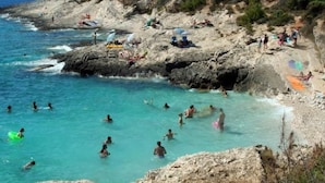 A rocky beach with clear turquoise water, people swimming and sunbathing, and a forested hillside in the background. - Wellness und Spa Villa Stella, in der Nähe von Stränden - Meer by Interhome (Bokordici)