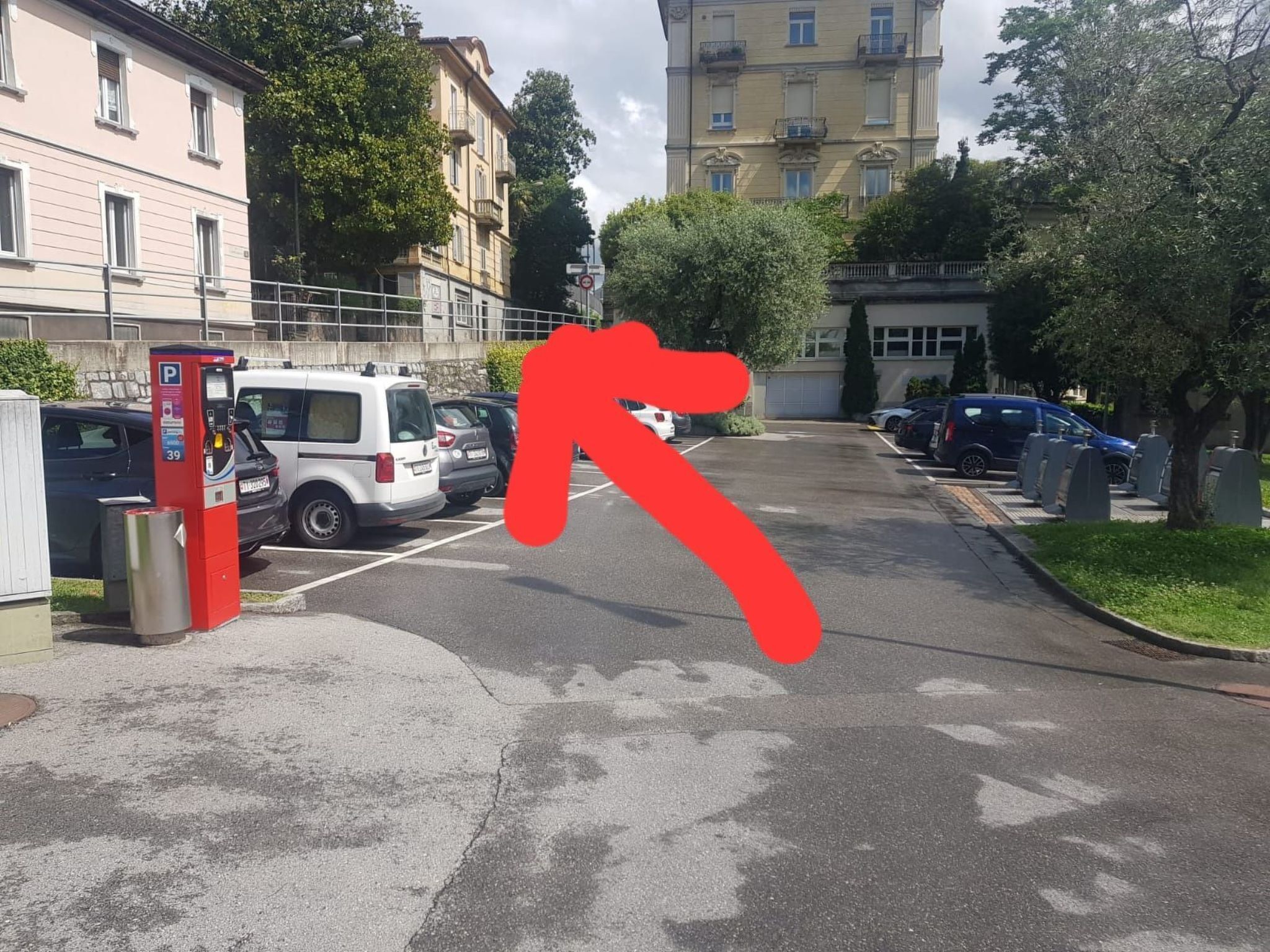 Um estacionamento com um parquímetro vermelho, uma van branca e vários carros estacionados.