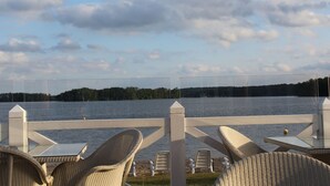 A lakeside patio with wicker chairs and tables, a white fence, and a view of the lake and forested area. - Appartement am See by Interhome (Bad Saarow)