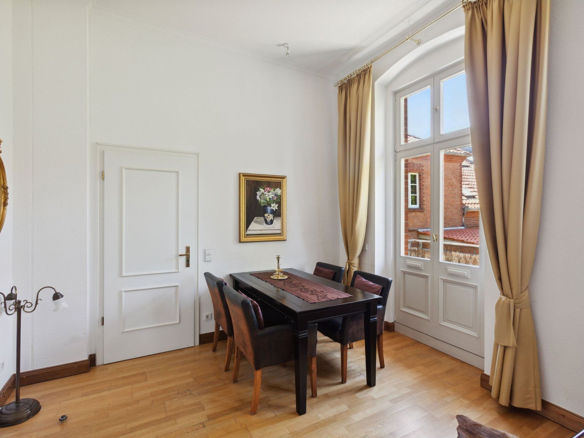 A dining room with a wooden table and chairs, a framed picture on the wall, and large windows with curtains.