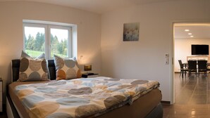 Un dormitorio con una cama grande, una ventana con vistas a árboles, un cuadro en la pared y una zona de comedor con sillas y un televisor.