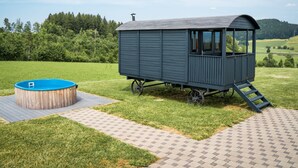 Una pequeña cabaña de madera azul oscuro con techo inclinado, una ventana y una puerta. Está ubicada en una zona pavimentada con un jacuzzi de madera cerca.
