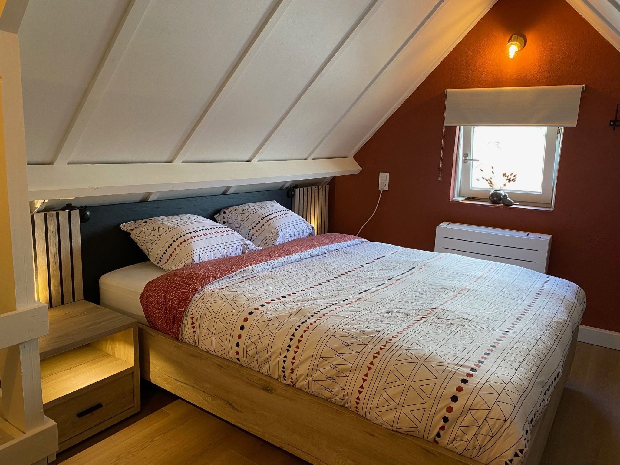 Ein Schlafzimmer mit geneigter Decke, einem Bett mit gemusterten Textilien, einem Nachttisch, einem Fenster mit Blick und einem Wandheizkörper.