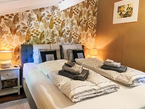 Ein Doppelbett mit gemusterten Betttextilien, zwei Nachttische mit Lampen und blumiger Tapete.