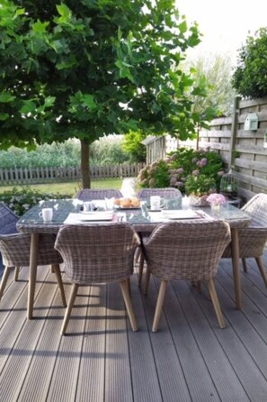 Un espace repas extérieur avec une table en verre et des chaises en osier, entouré d’une terrasse en bois et d’un jardin.