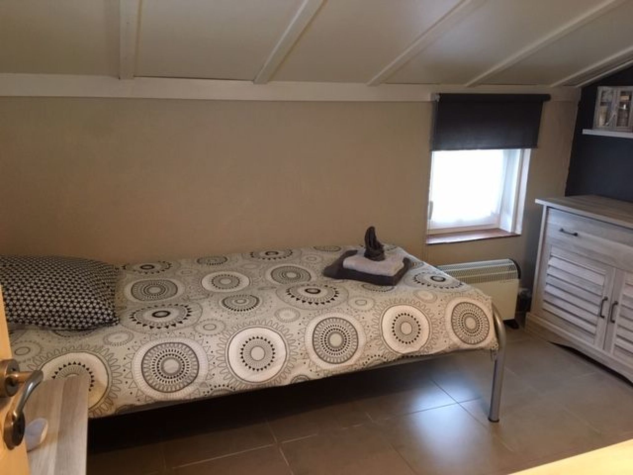 Une petite chambre avec un lit simple, une couvre-lit à motifs, une table de chevet avec une lampe, une fenêtre avec un store et une armoire intégrée.
