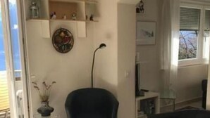Ein Zimmer mit Fliesenboden, einem schwarzen Stuhl, einem kleinen Tisch, einem Regal mit Dekoration und einem Fenster mit Jalousien.