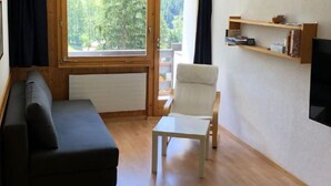 Una sala de estar con sofá, sillón, una mesita, televisión y ventana con vista a árboles.