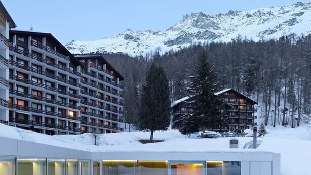 Un hotel moderno con varios pisos y balcones, rodeado de montañas nevadas y árboles.