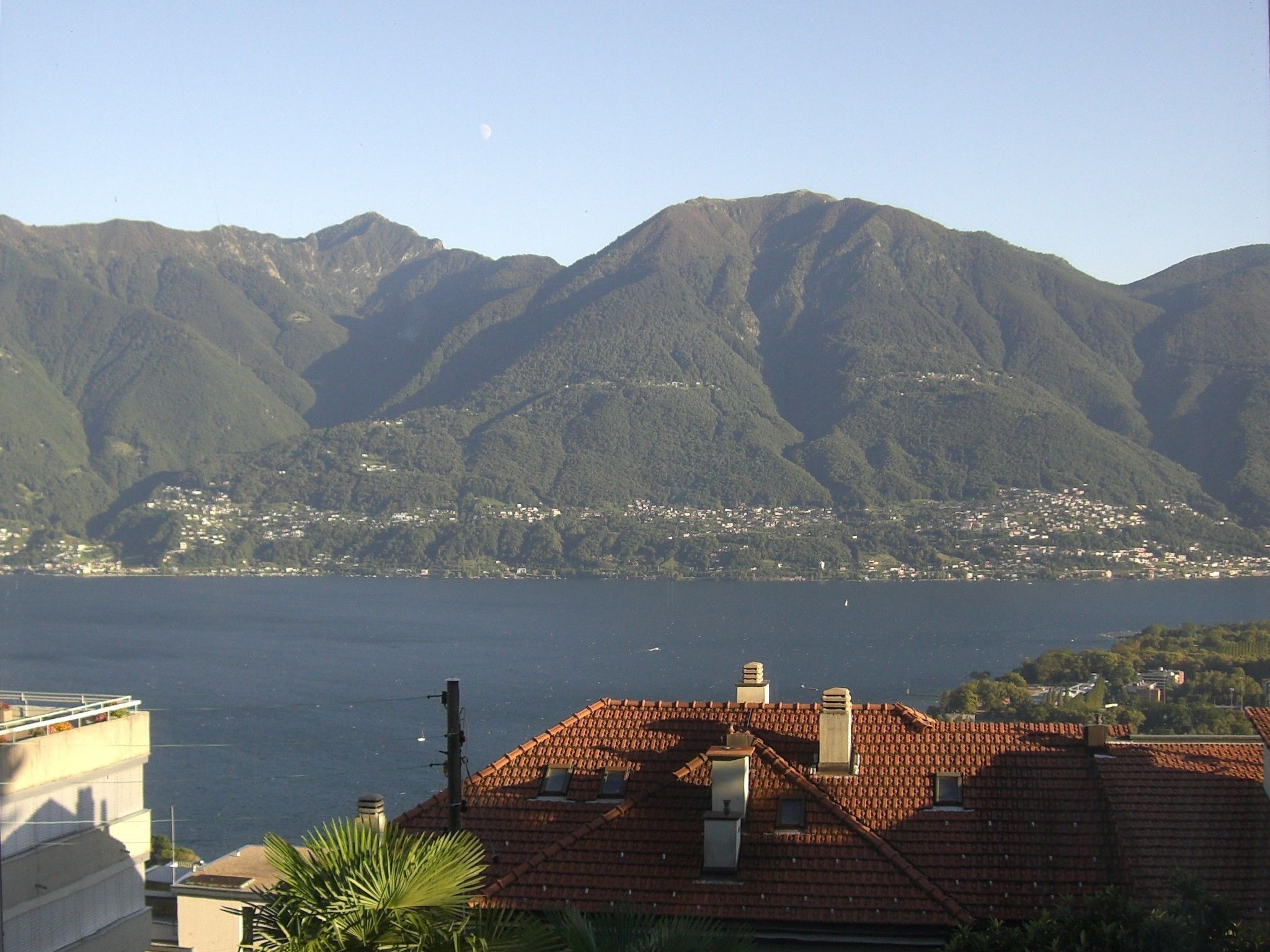 Une ville côtière aux bâtiments aux toits de tuiles rouges, bordée d’une étendue d’eau, avec des montagnes en toile de fond.