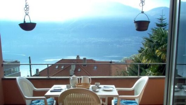 Ein Balkon mit Tisch und St ühlen, der auf eine Berglandschaft und ein Gewässer blickt.