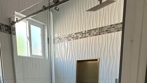 Un baño moderno con ducha de vidrio, paredes de azulejos blancos y un pequeño espejo con marco dorado.