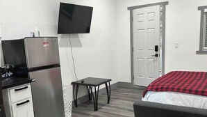 Una habitación compacta con cama, refrigerador, una mesita y televisor montado en la pared.