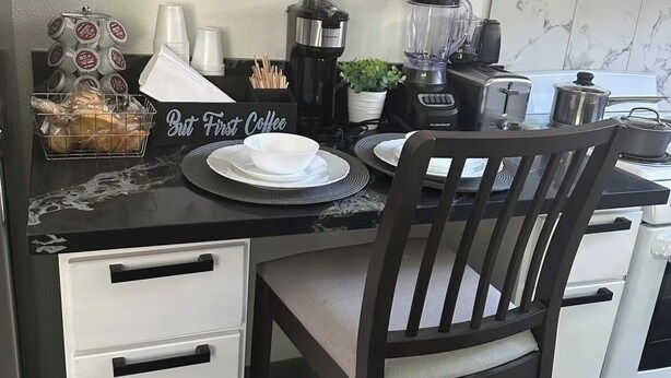 Una cocina compacta con armarios blancos, encimera negra y una silla de madera. Hay tazas de café, una cafetera y una licuadora sobre la encimera.