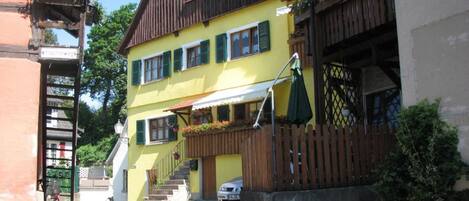 A two-story building with a yellow facade, green shutters, and a wooden staircase leading to the entrance.